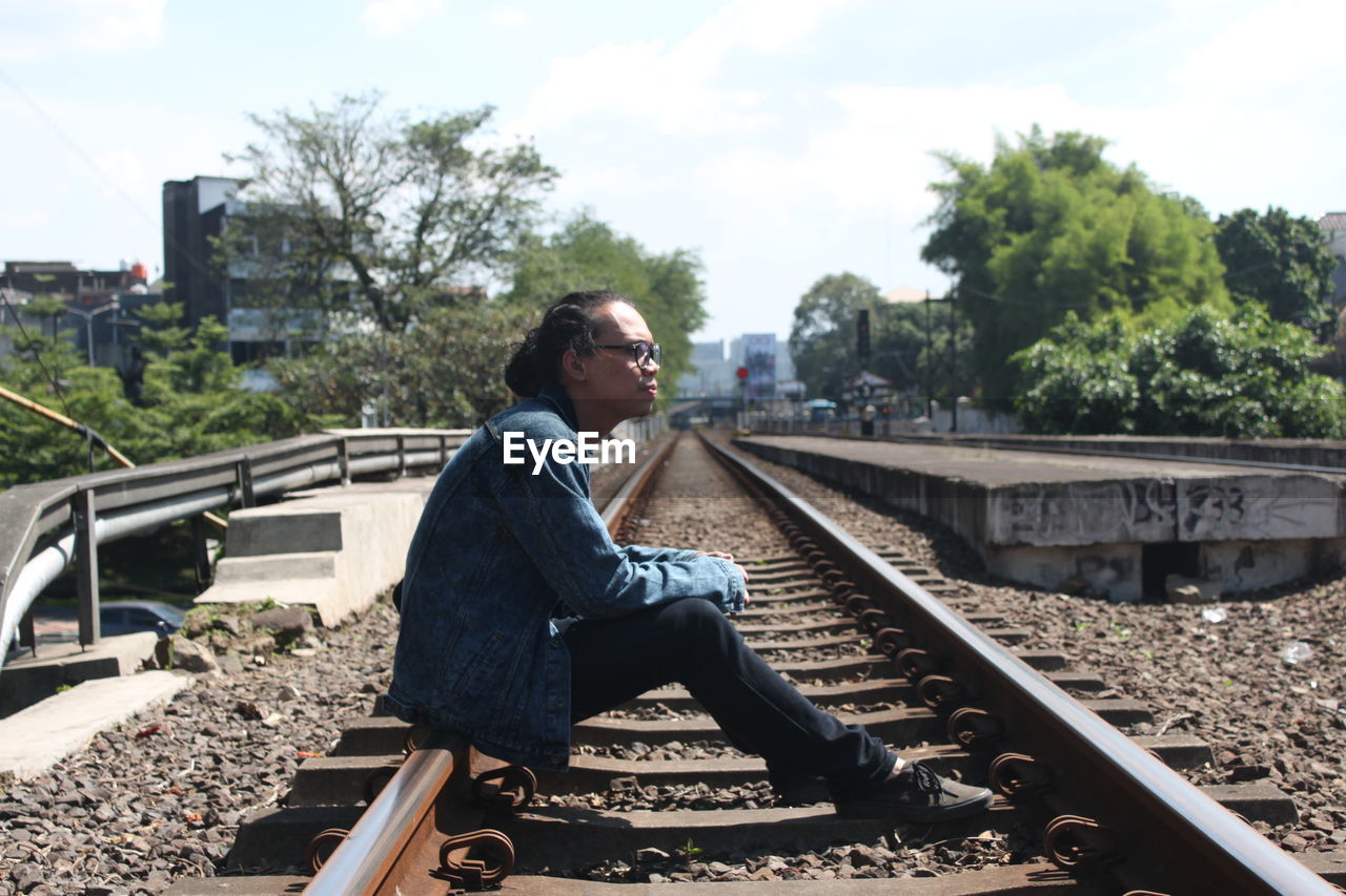 Side view of young man sitting on railroad track against sky