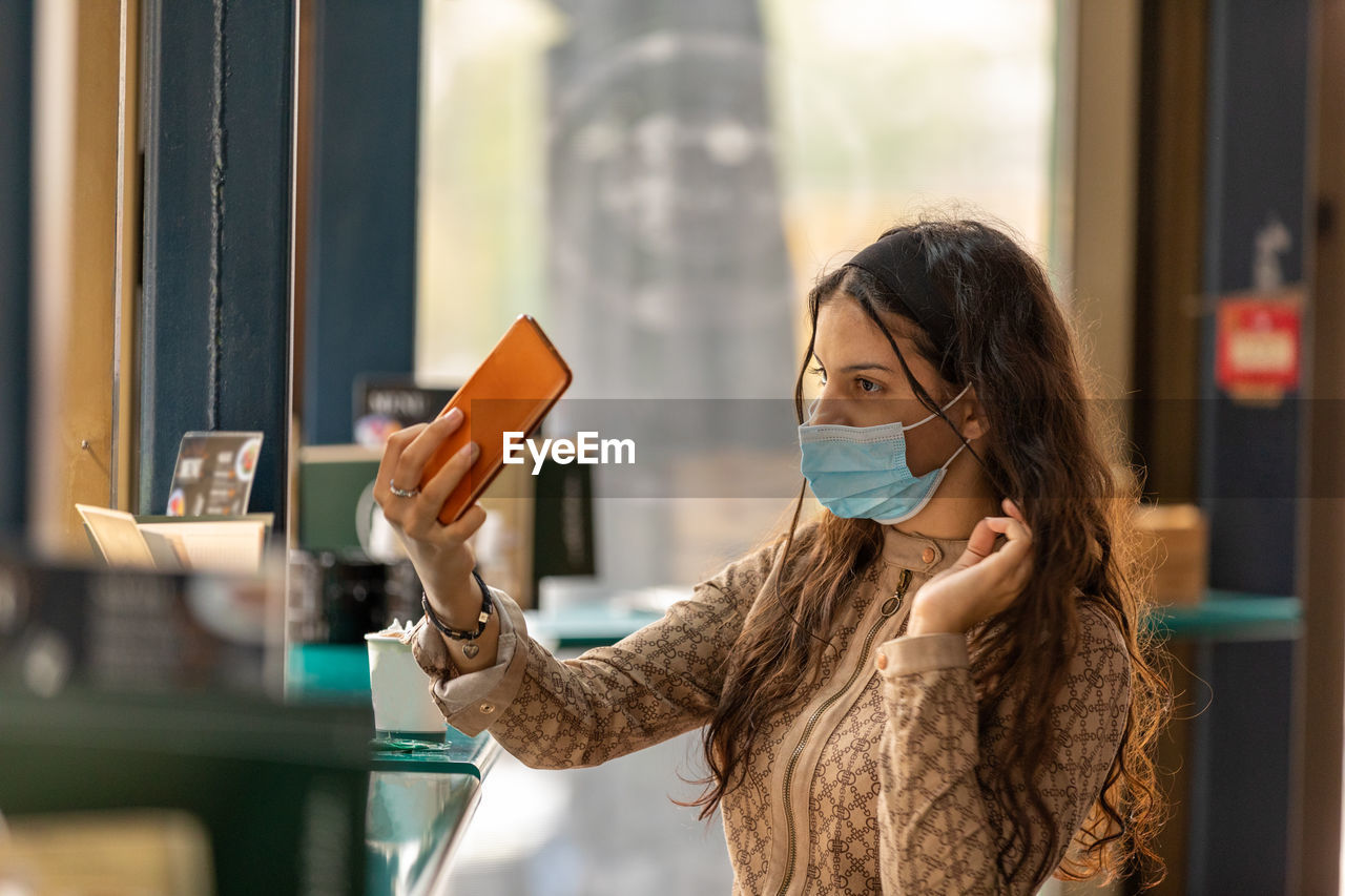 Cute teen wearing medical mask sitting in cafe touching and combing hair using smartphone as mirror