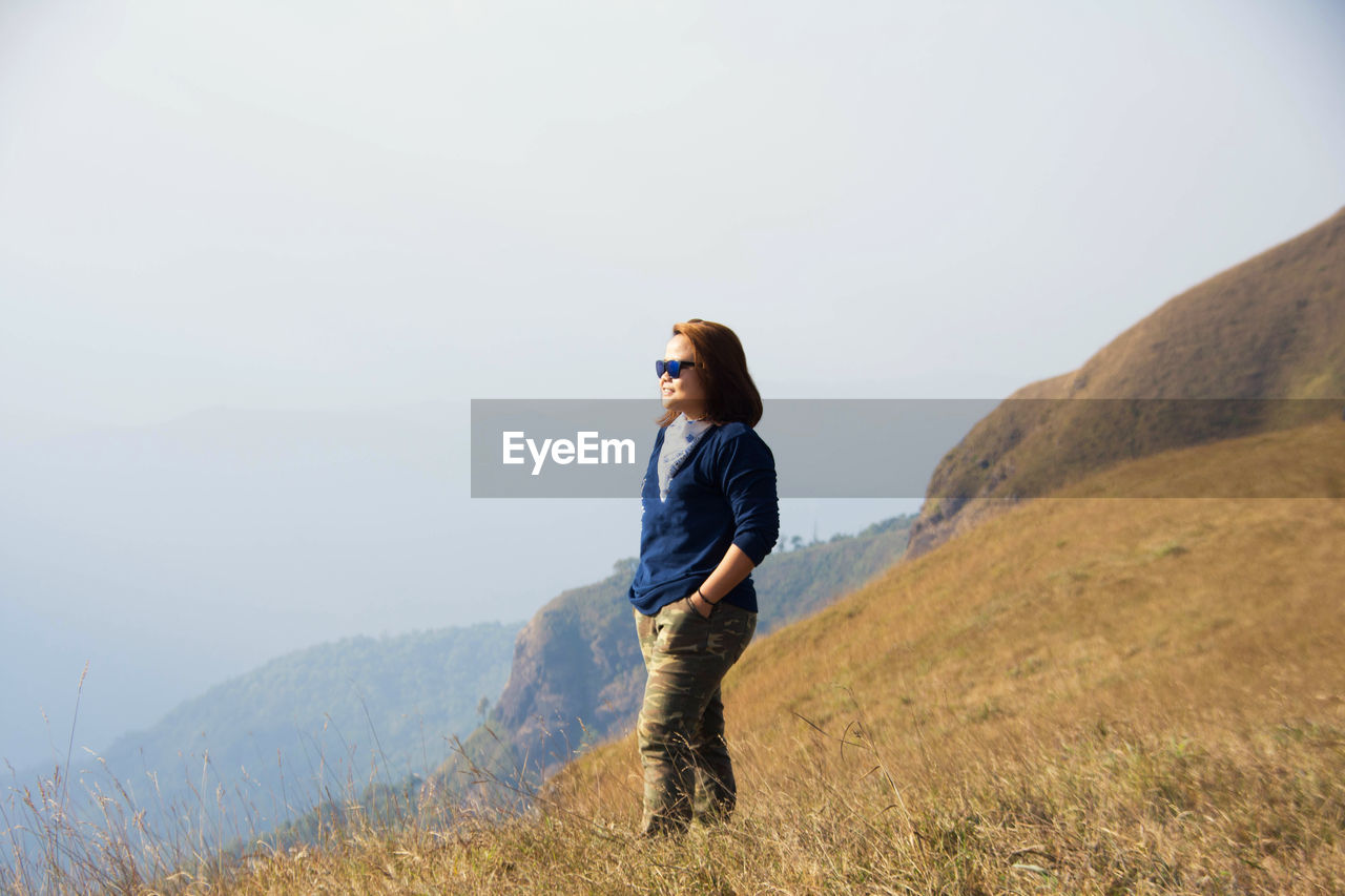 Woman with hands in pocket standing on hill against sky during sunny day