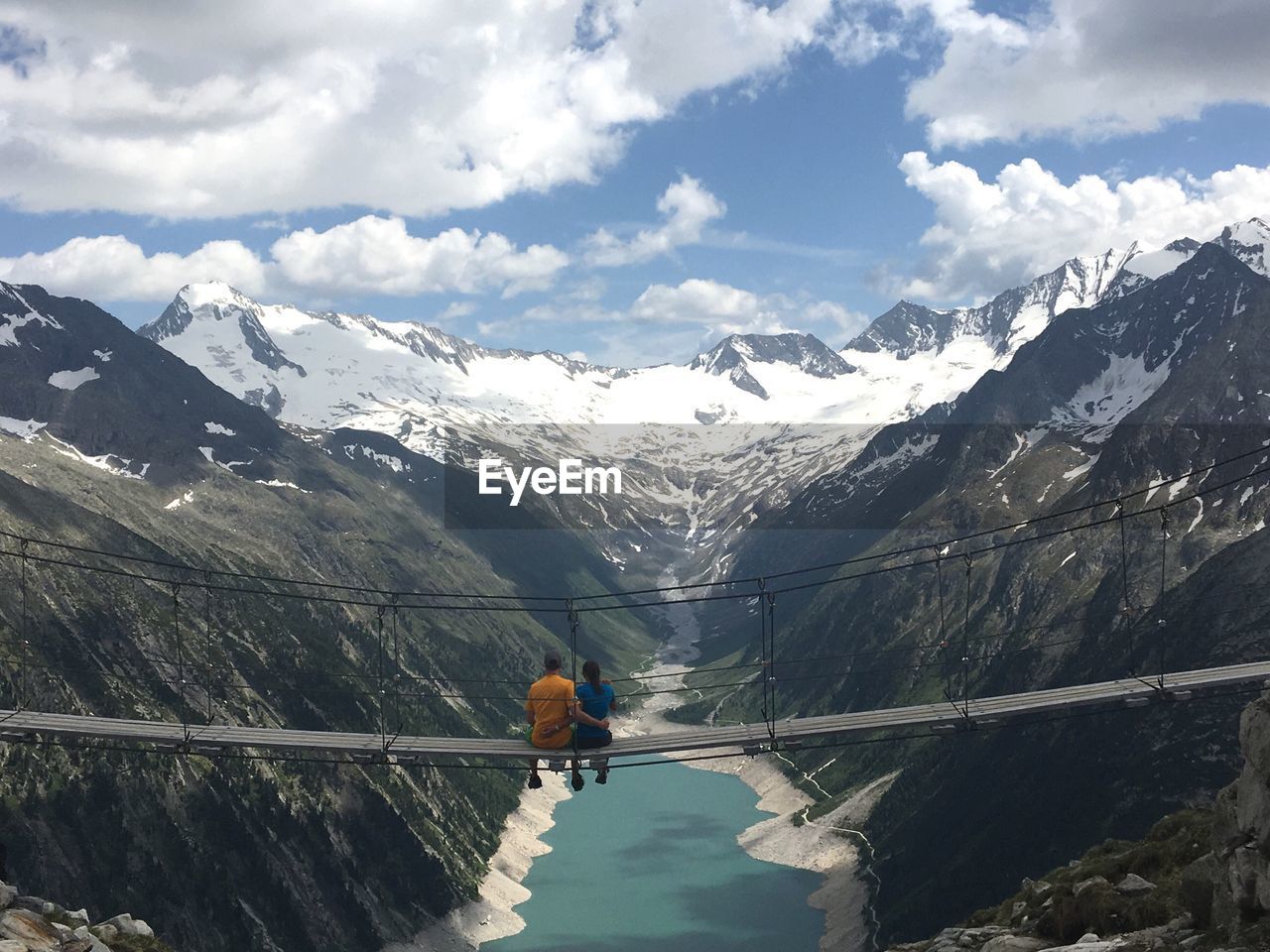 Rear view of couple looking at snowcapped mountains while sitting on footbridge over river against cloudy sky