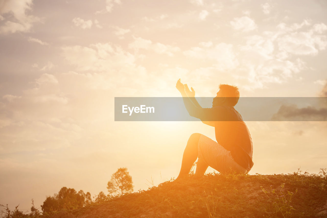 Silhouette man praying while sitting on land against sky during sunset