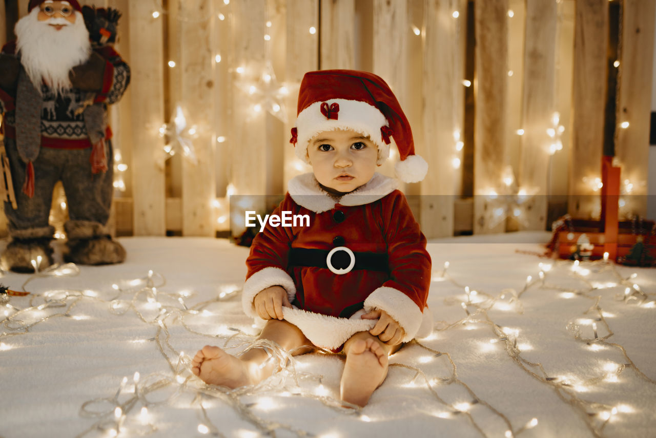 A baby girl sits looking at the camera dressed as a santa claus.