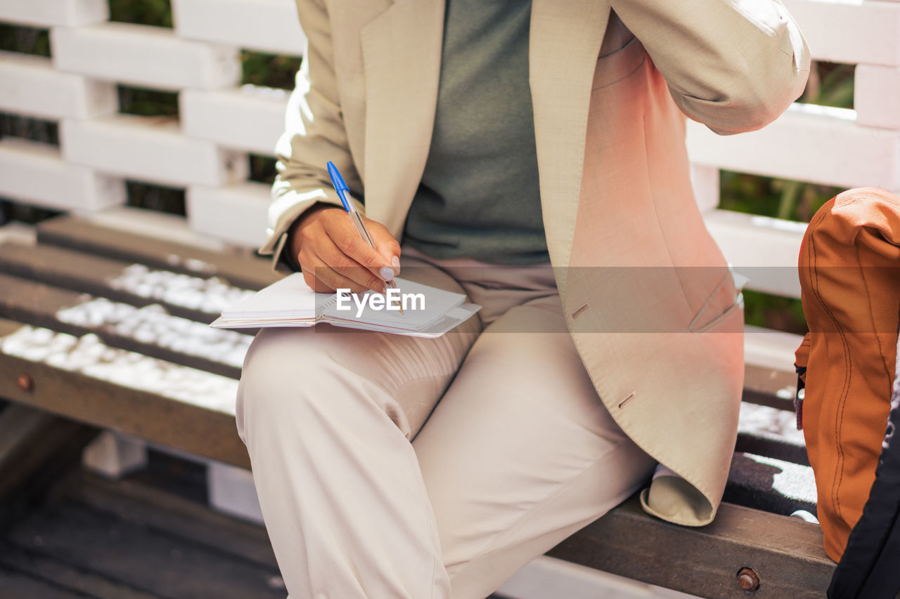 Cropped unrecognizable entrepreneur in suit sitting on bench and writing in planner while working remotely on sunny day in street restaurant