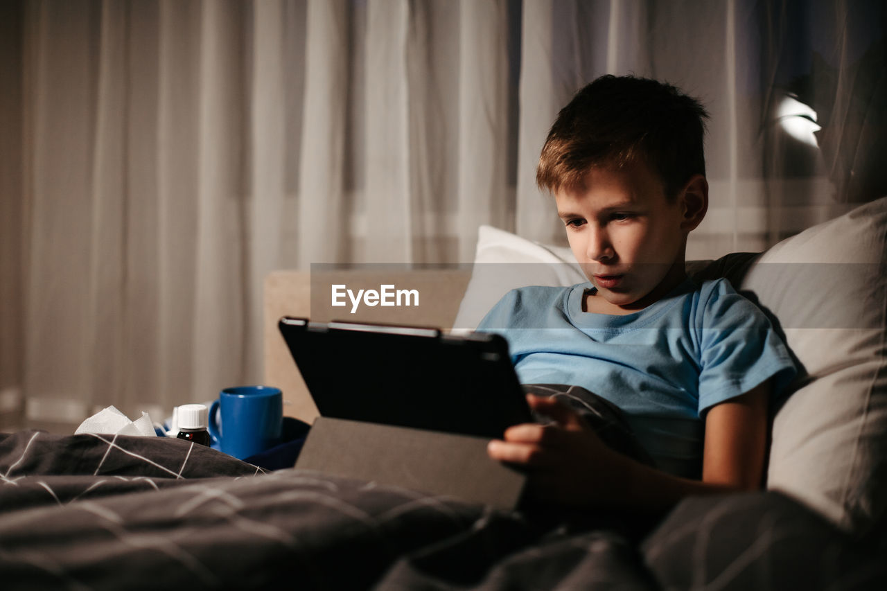 Little boy using tablet while laying in bed in living room. medicines and thermometer nearby. 