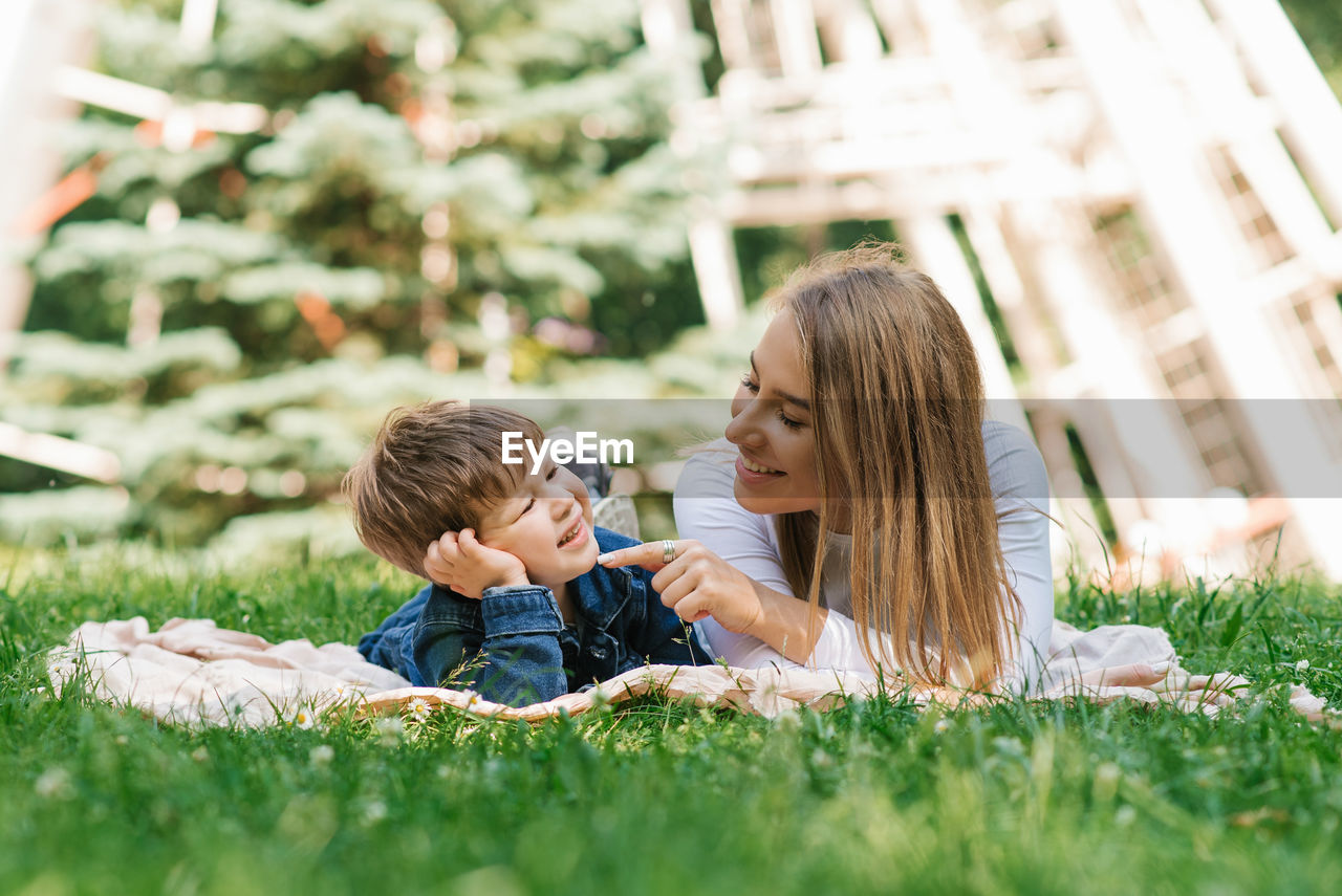 Young caucasian mother and her young son lie on a blanket on the grass in the park in summer