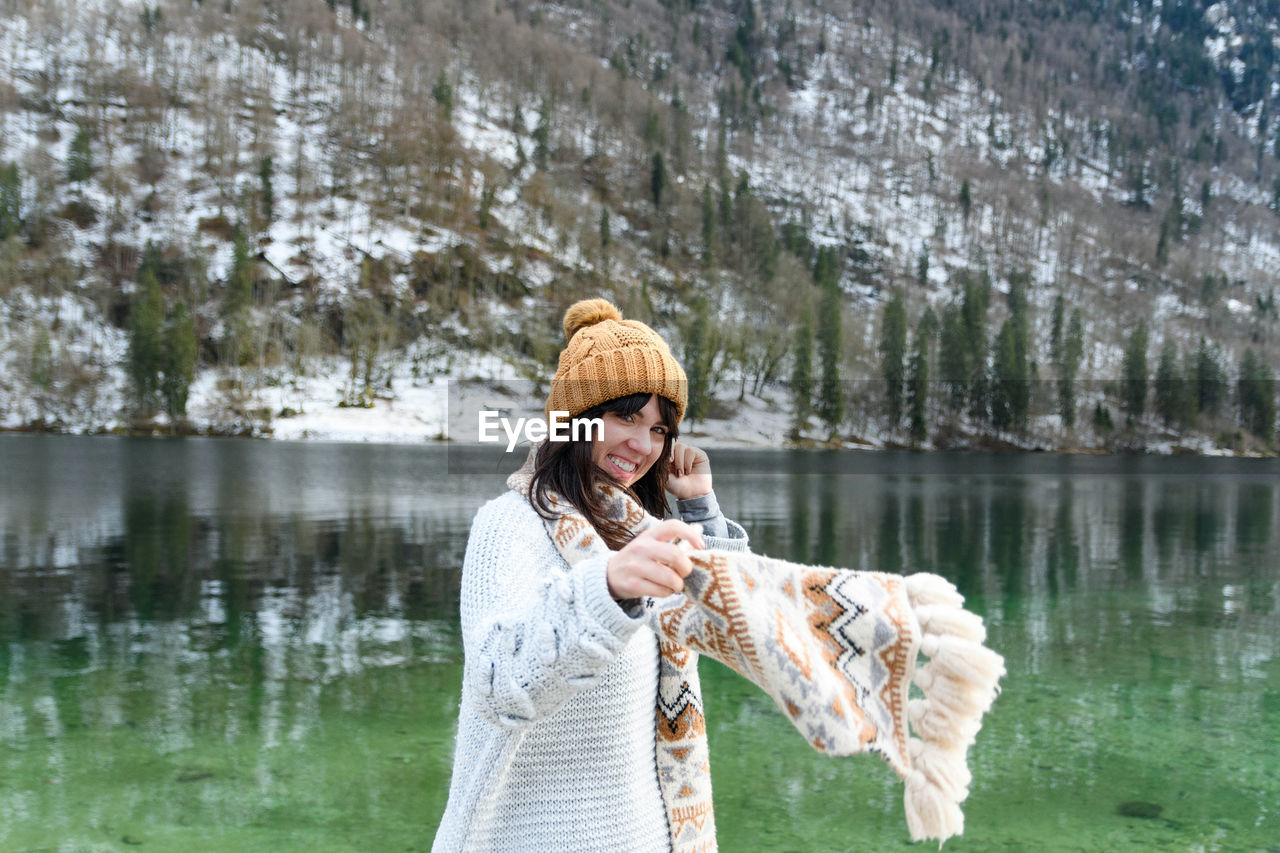 Young woman in winter sweater smiling, standing by a lake.