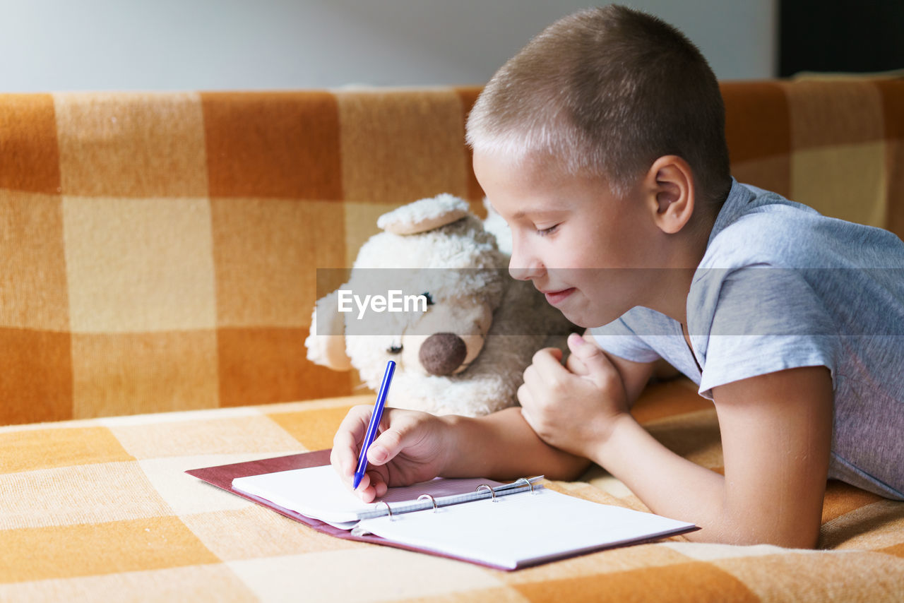 Caucasian boy lying on couch does his homework, writes with a pen in a notebook