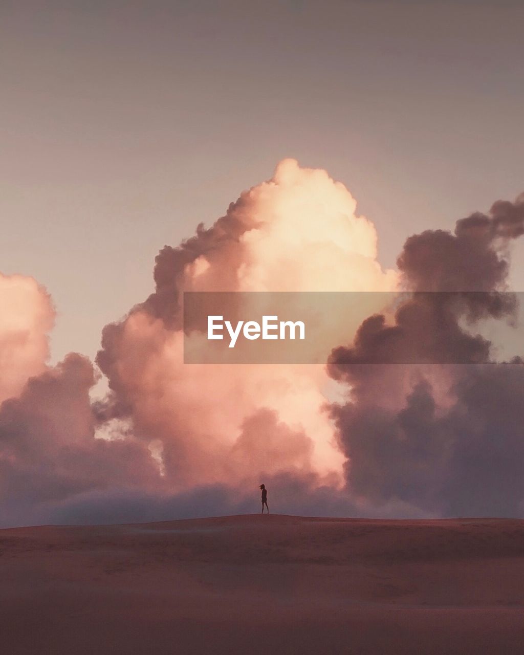 Distant view of silhouette woman standing on sand dune against sky during sunset