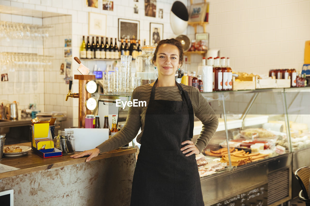 Portrait of confident young female owner standing with hand on hip at cafe