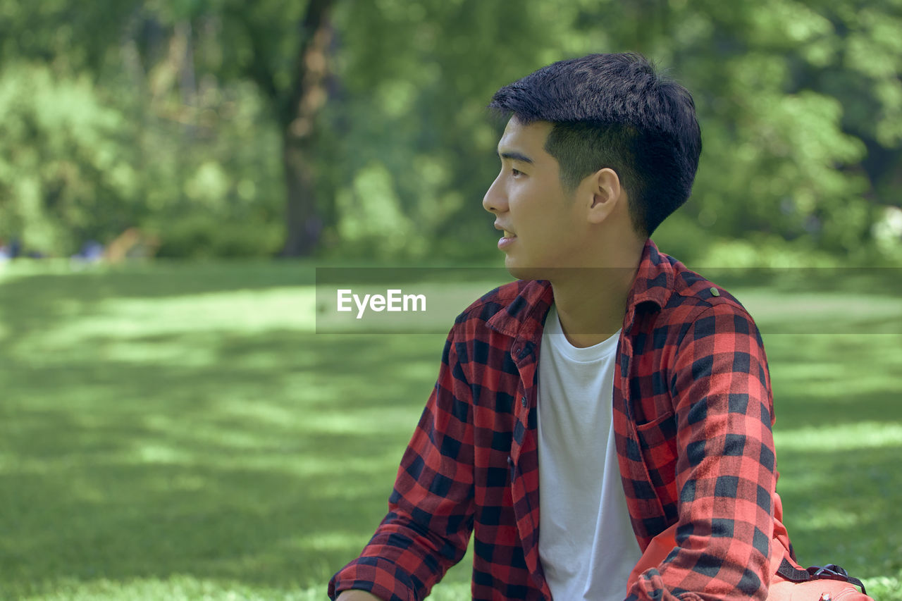 A handsome young asian man sits on the lawn in the summer at the park.