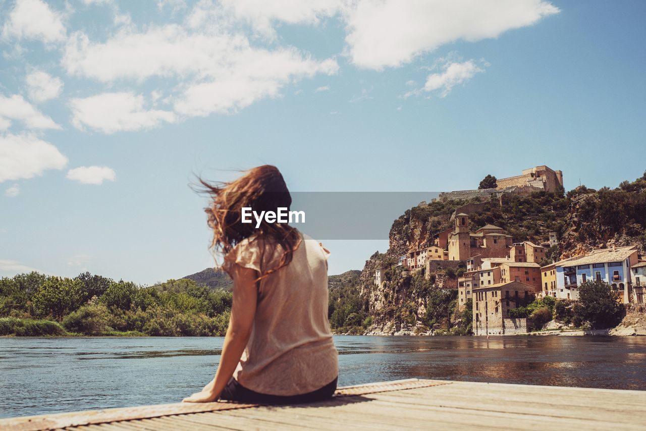 Rear view of woman with tousled hair sitting on pier by lake