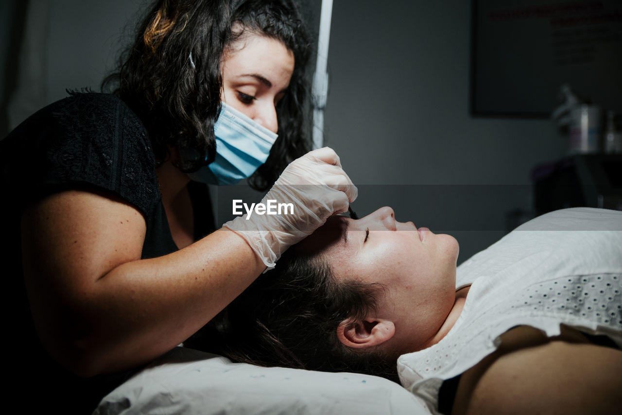 Side view of focused cosmetician in latex gloves curling eyelashes of female patient during lash lift procedure in salon