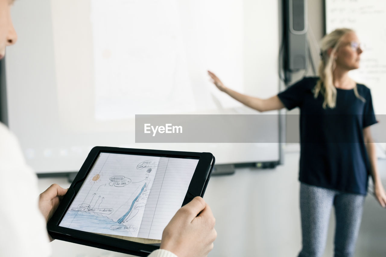 Cropped image of boy holding digital tablet with teacher explaining in classroom