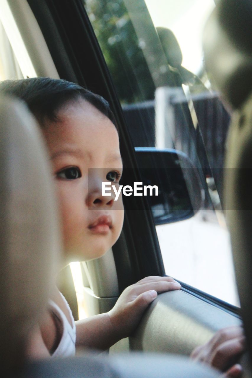 Close-up of cute baby boy looking through window in car