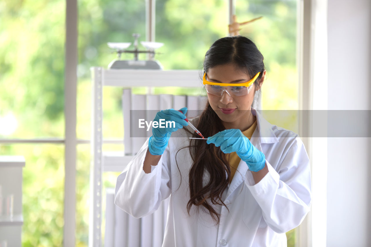 PORTRAIT OF YOUNG WOMAN HOLDING EYEGLASSES WHILE STANDING AGAINST WINDOW