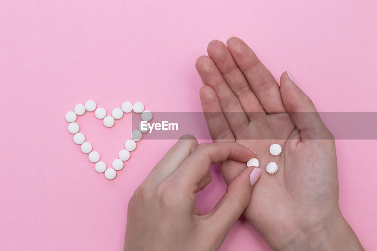 Cropped hands holding pills by heart shape on pink background