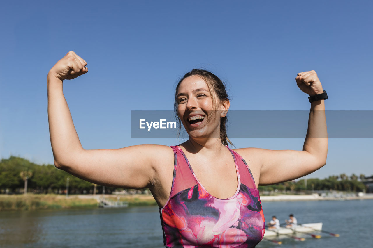 Sportswoman laughing while flexing muscles at riverbank