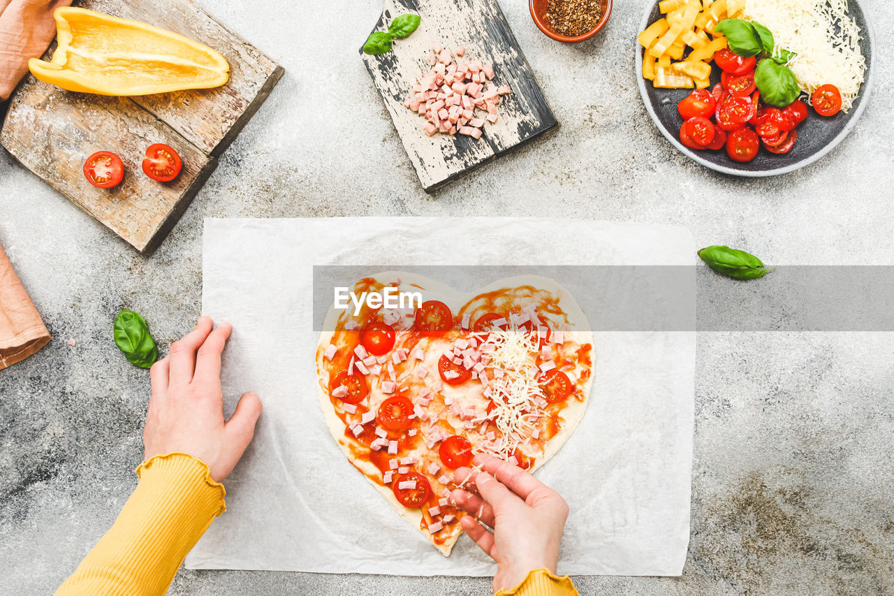 Hands of caucasian teenage girl lay down sliced cherry tomato on heart-shaped pizza dough.