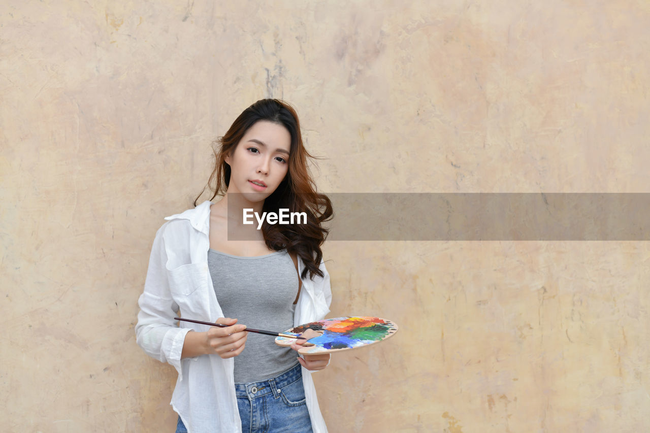 Portrait of young woman holding paintbrush standing against brick wall