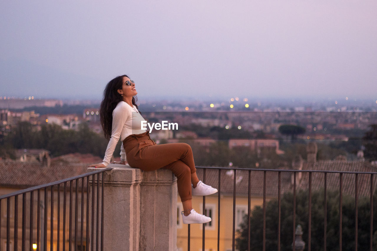 Full length of woman sitting by railing against cityscape