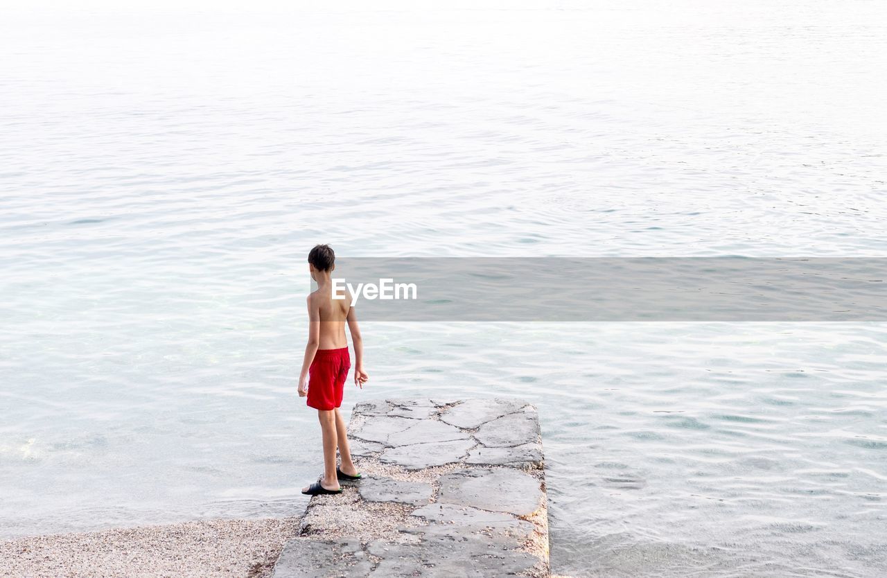 Rear view of shirtless teenage boy standing on pier at beach