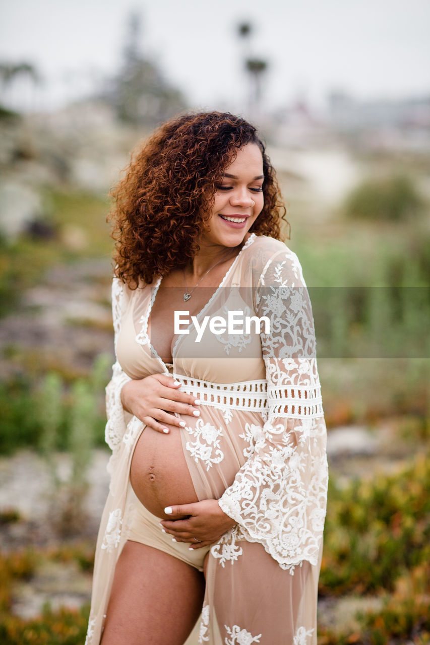 Young pregnant woman posing & smiling at beach in sheer dress