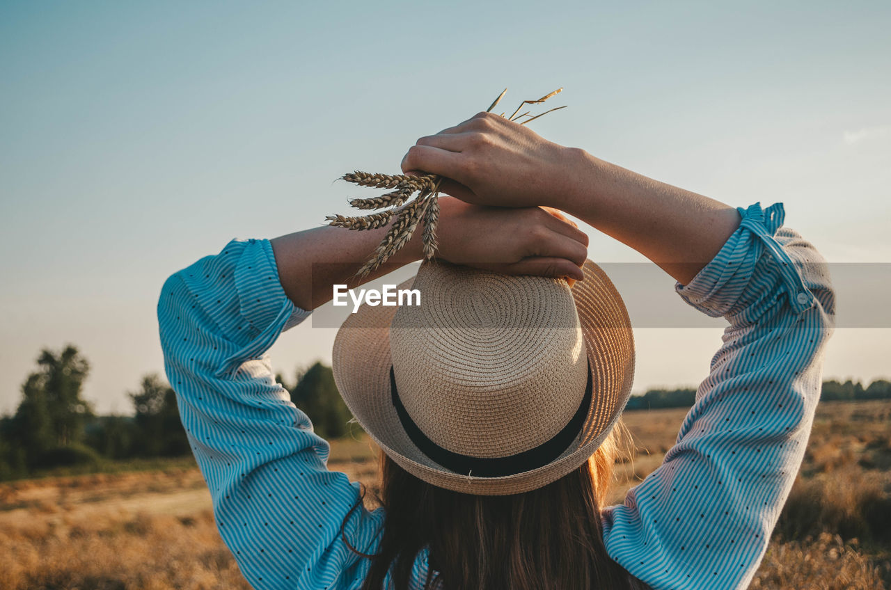 Rear view of woman wearing hat holding wheat against sky at sunset