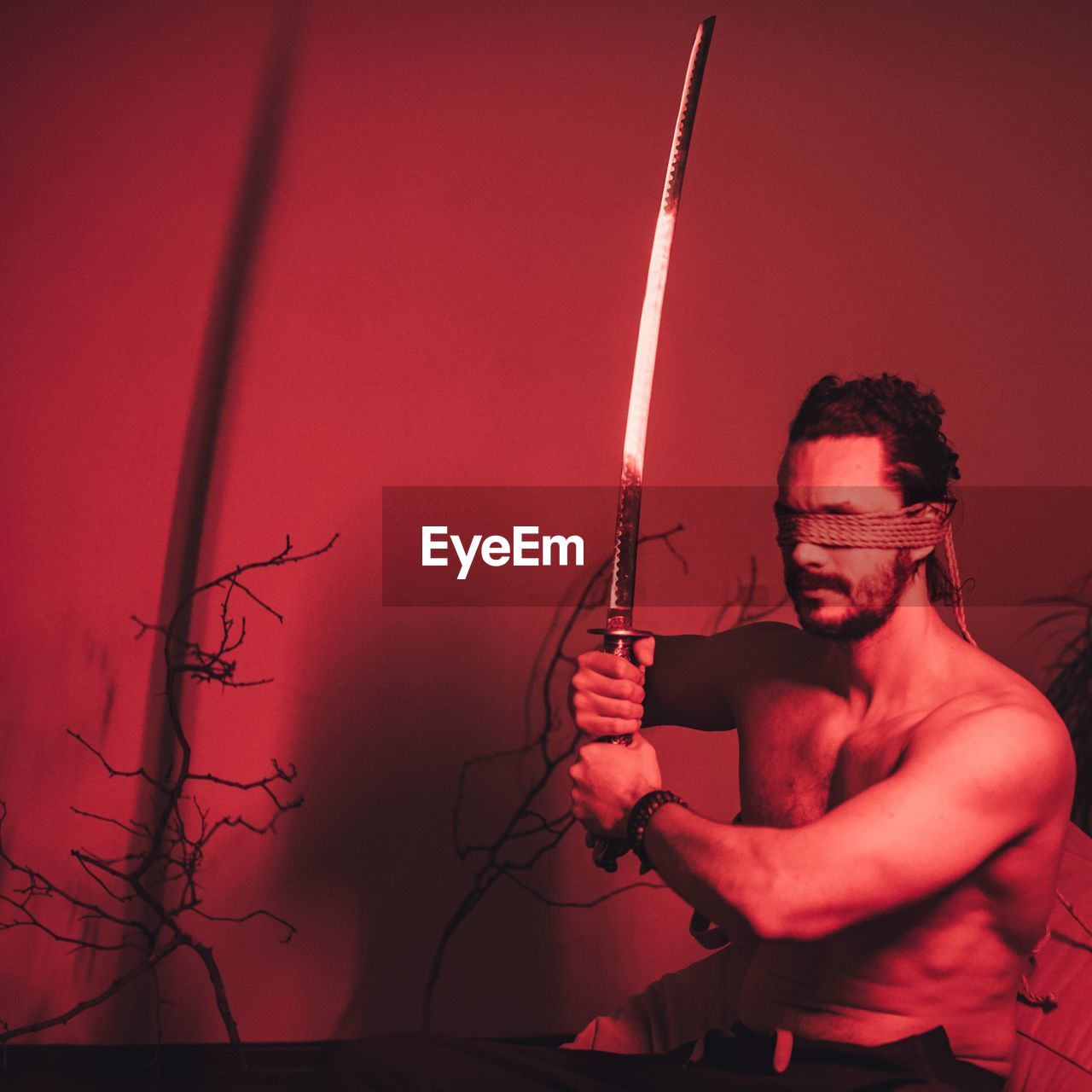 Shirtless man with blindfolds holding sword in illuminated room