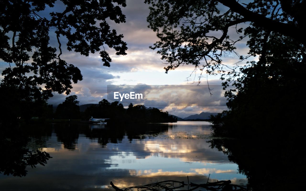 REFLECTION OF SILHOUETTE TREES IN LAKE AGAINST SKY DURING SUNSET