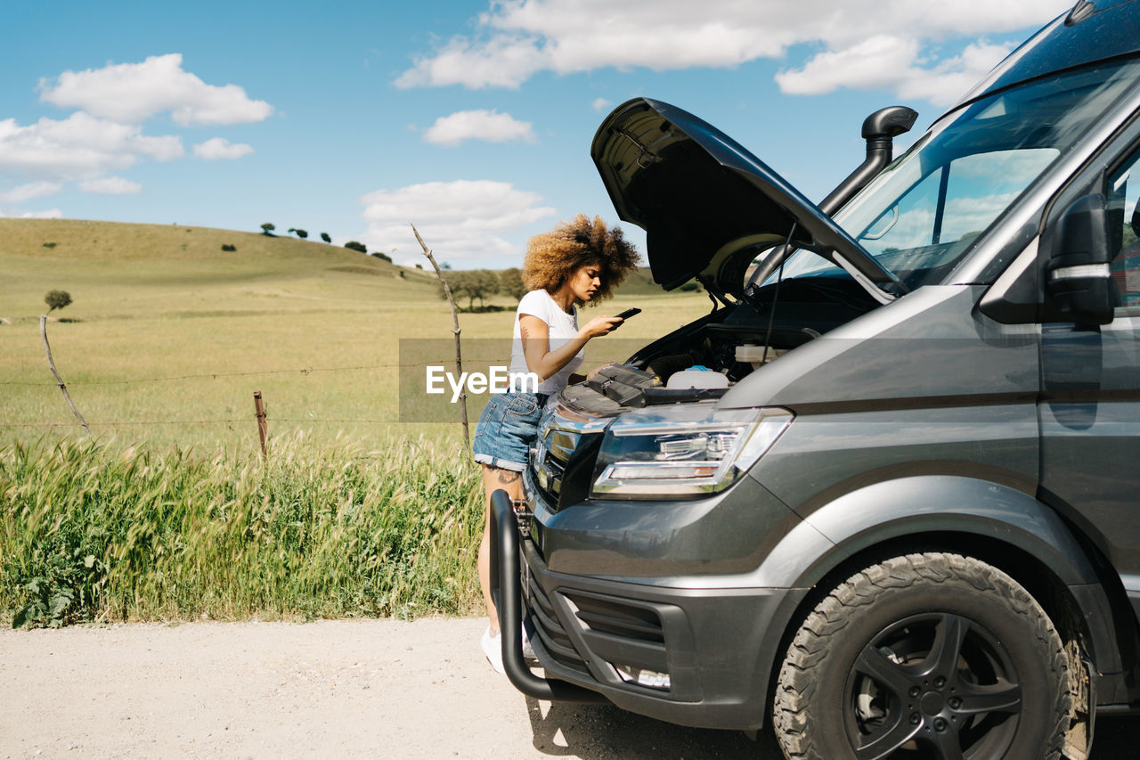 Young ethnic female with afro hair calling to repair service on smartphone while standing near camper van with open hood on countryside road in summer day