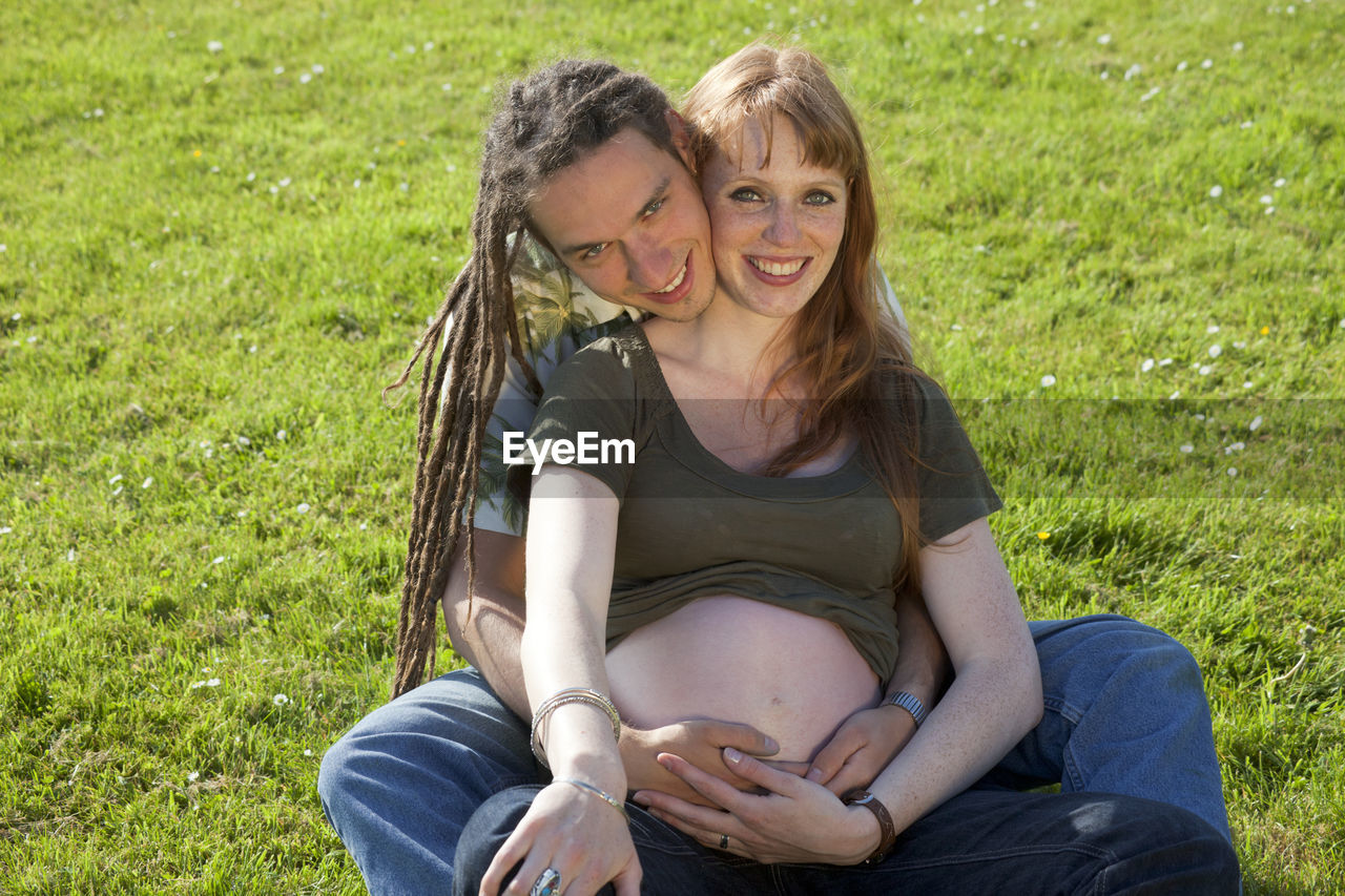 Portrait of happy man sitting with pregnant woman on grassy field