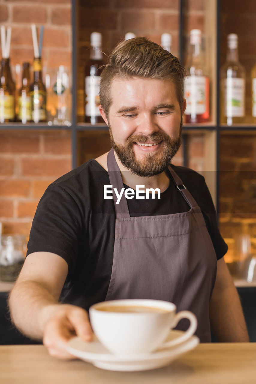Bearded male barista in apron serving cup of coffee while working at bar table in coffee shop