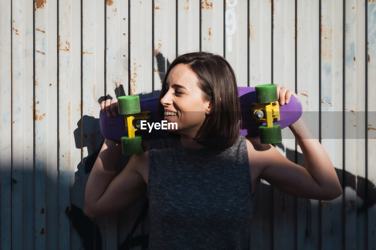 Smiling young woman holding skateboard while standing against wall