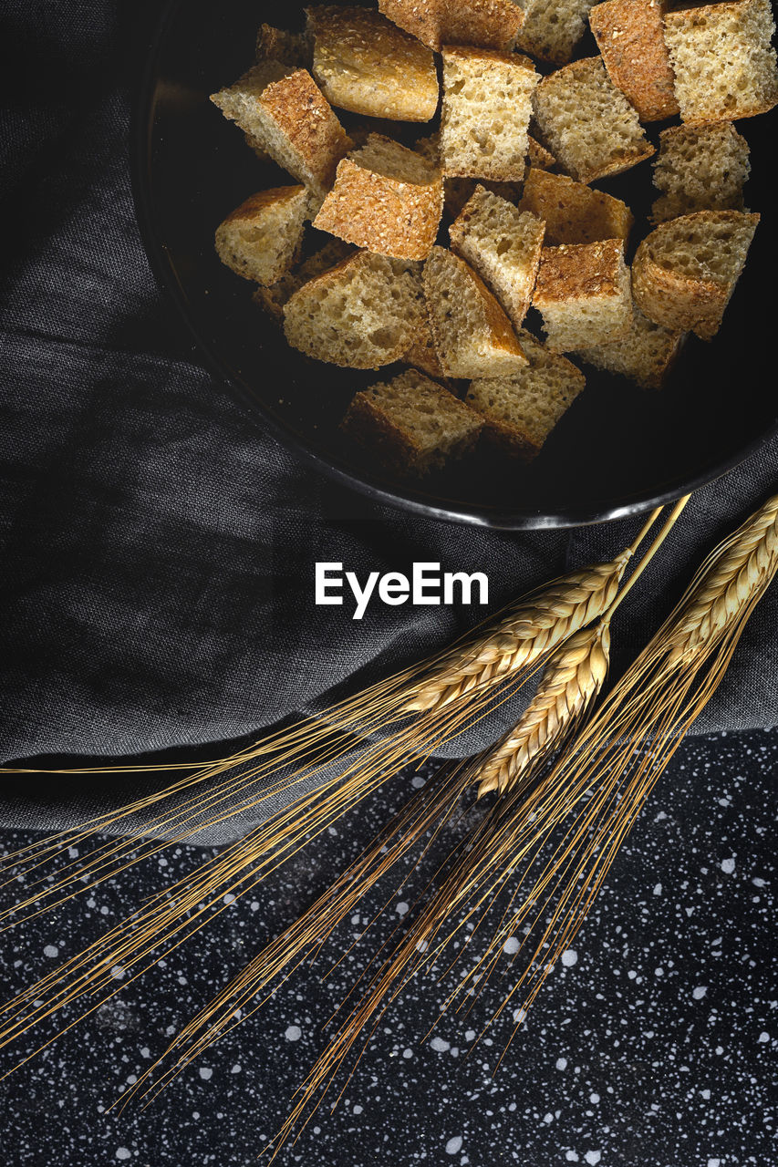 Pieces of crispy bread in bowl near wheat spikes on black textile in room