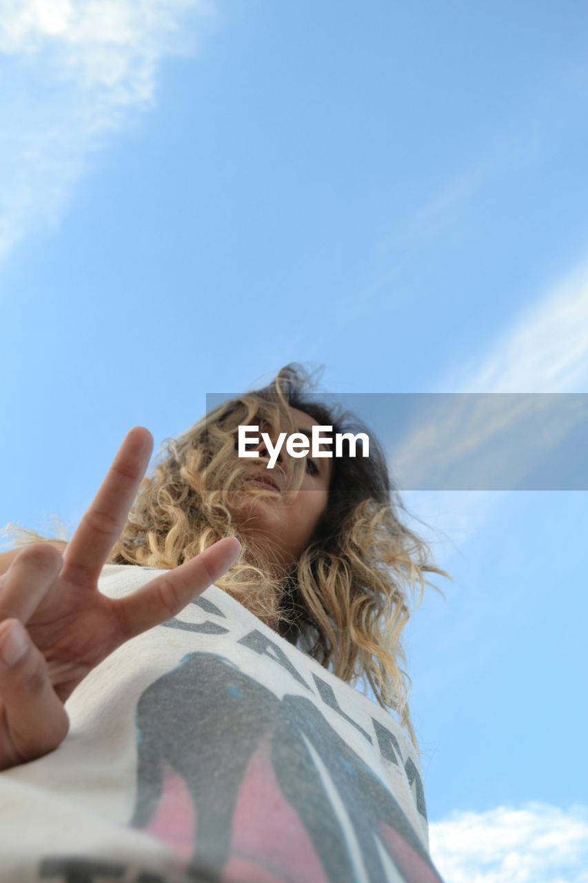 Low angle portrait of young woman showing peace sign while standing against blue sky