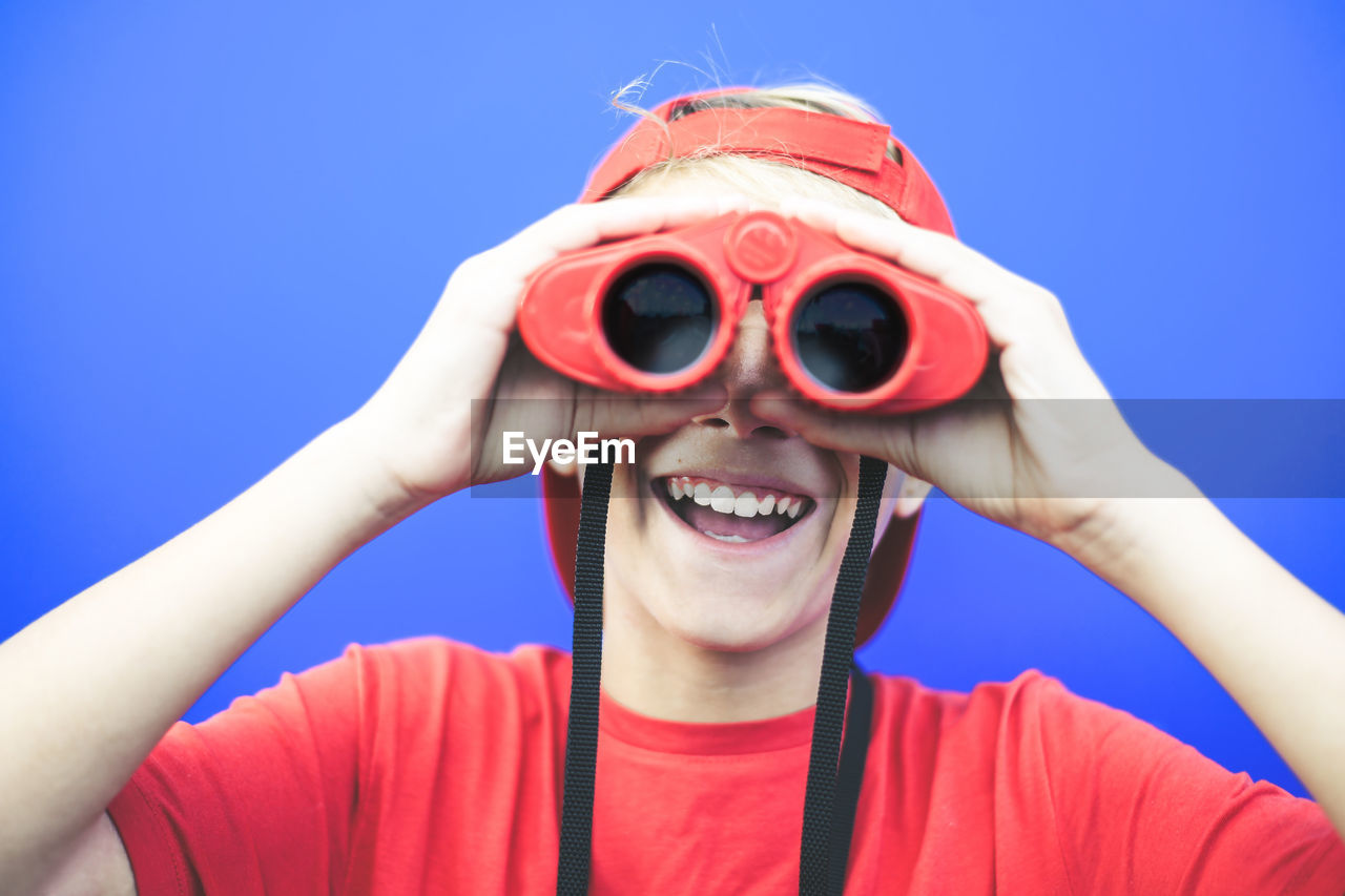 Close-up of boy looking through binoculars while standing against blue background