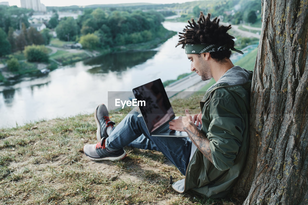 Side view of young man using laptop while sitting on grass by lake