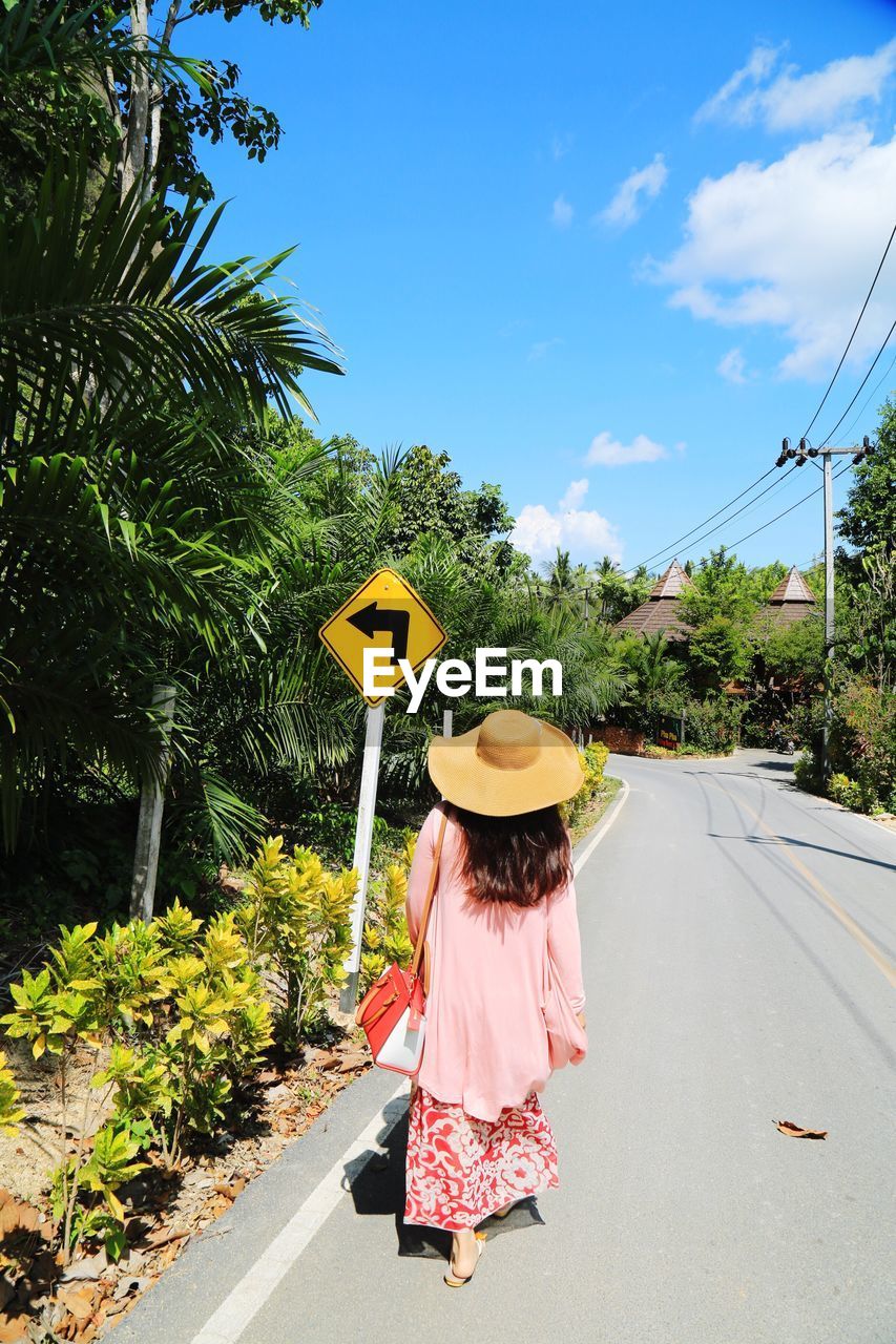 Rear view of woman walking on road amidst trees against sky