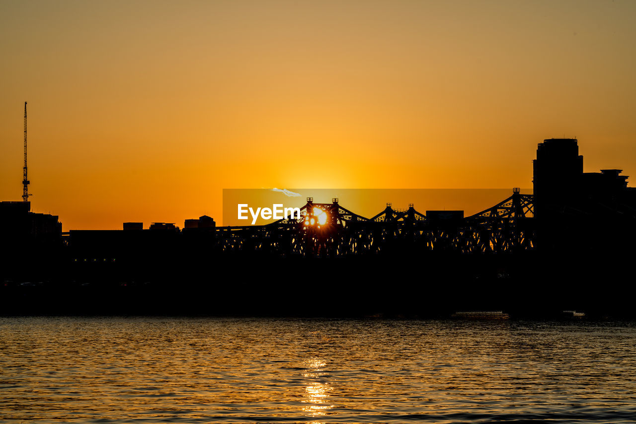 Silhouette buildings by river against sky during sunset