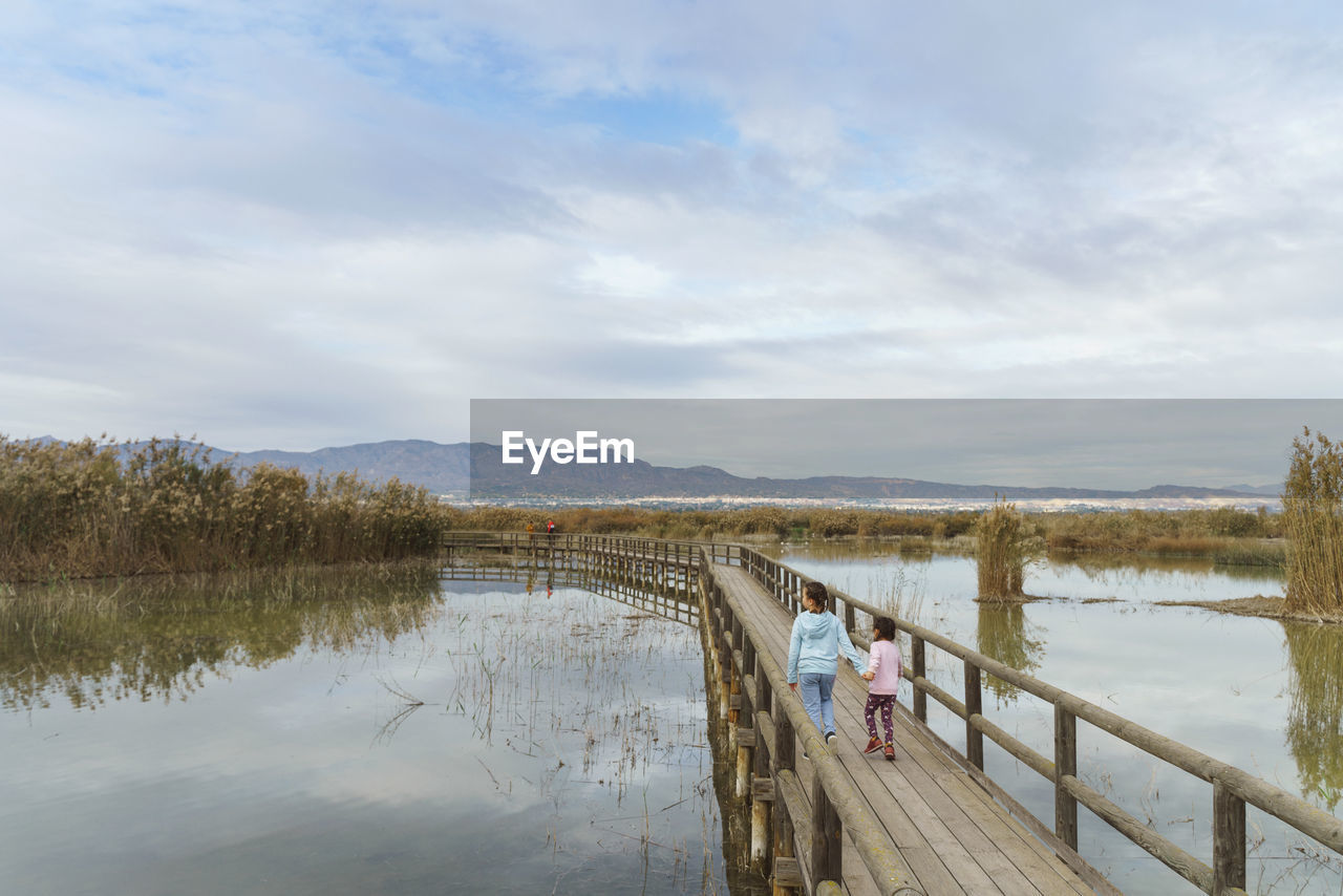 Pier over lake against sky and two girls running on a bridge 