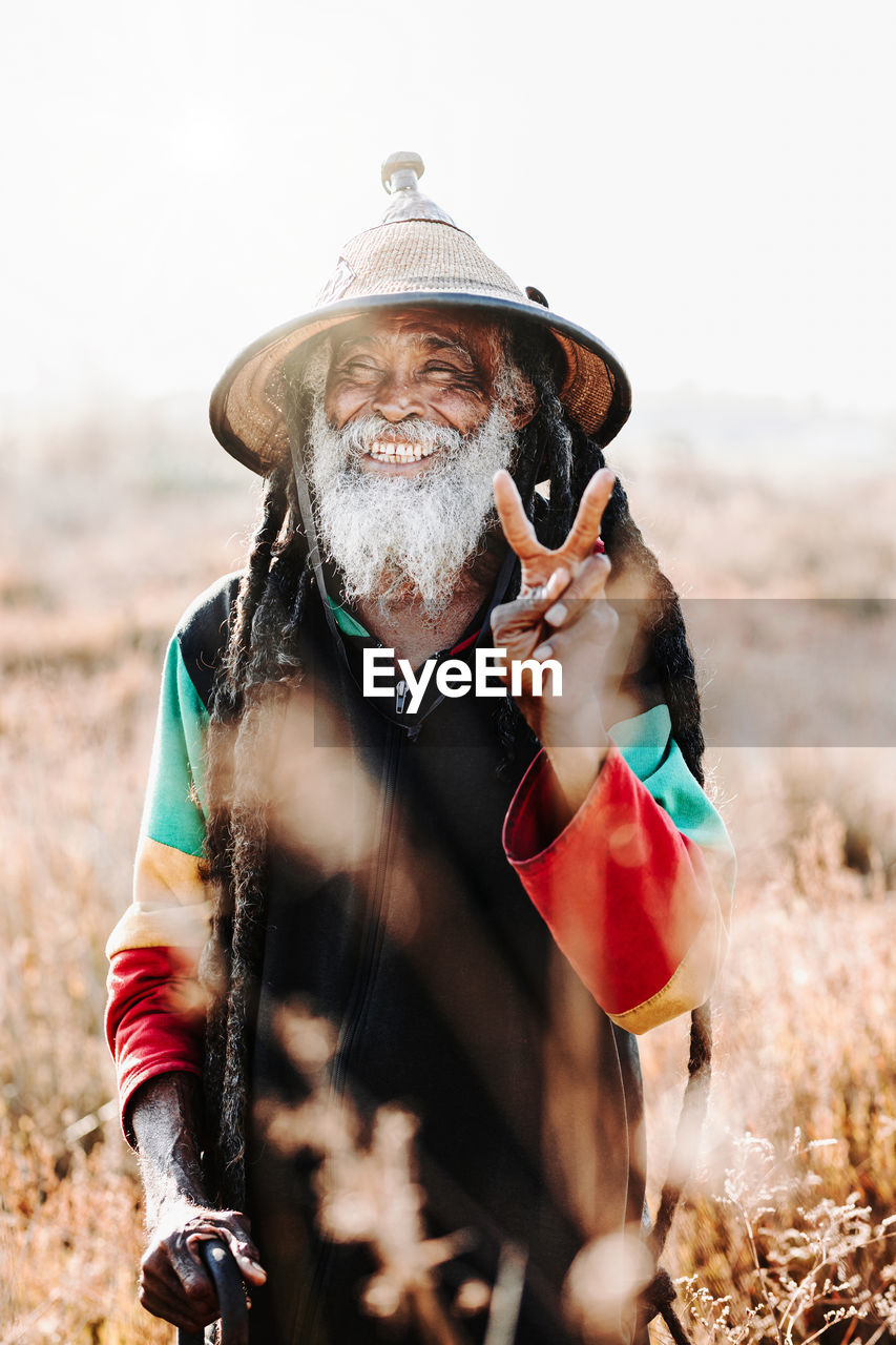 Cheerful old ethnic rastafari with dreadlocks looking at the camera in standing in a dry meadow in the nature
