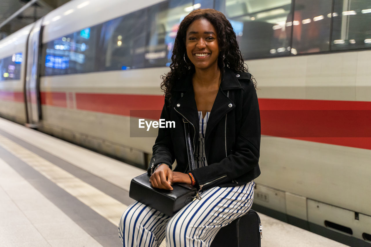 Portrait of smiling young woman sitting against train at railroad station platform