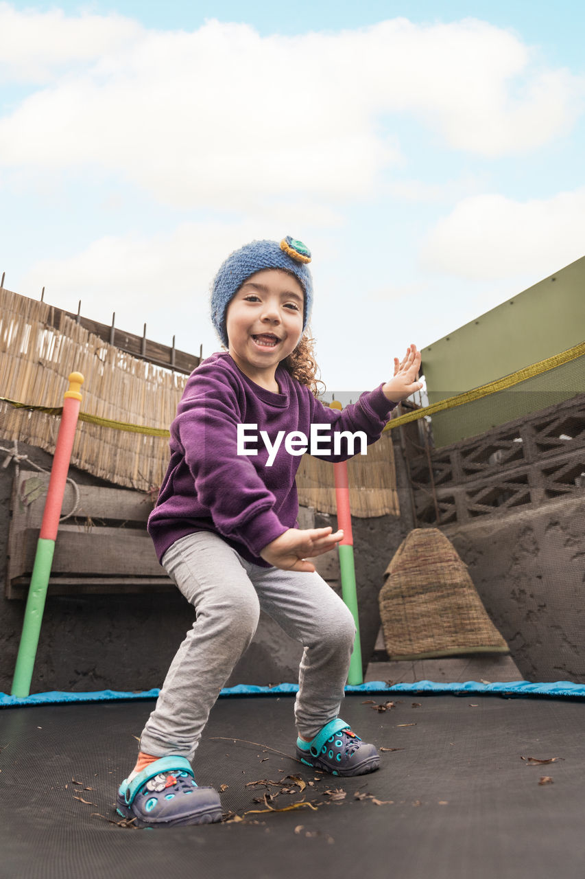 Full body of positive ethnic child in headband looking at camera with smile while jumping on trampoline and having fun in countryside