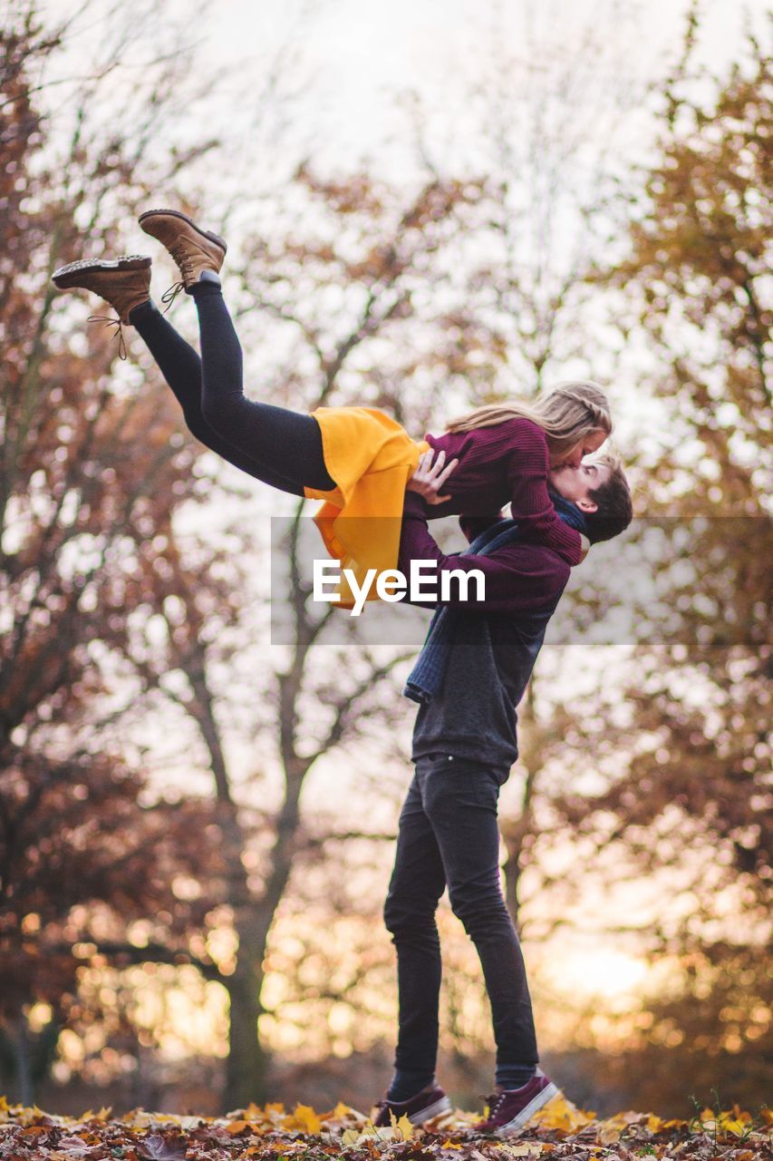 Young man carrying woman and kissing while standing against trees