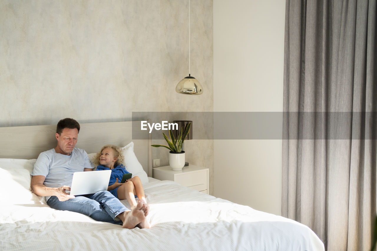 Smiling daughter with mobile phone by father using laptop on bed