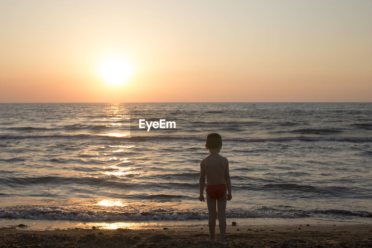 Rear view of shirtless boy standing at beach against clear sky during sunset