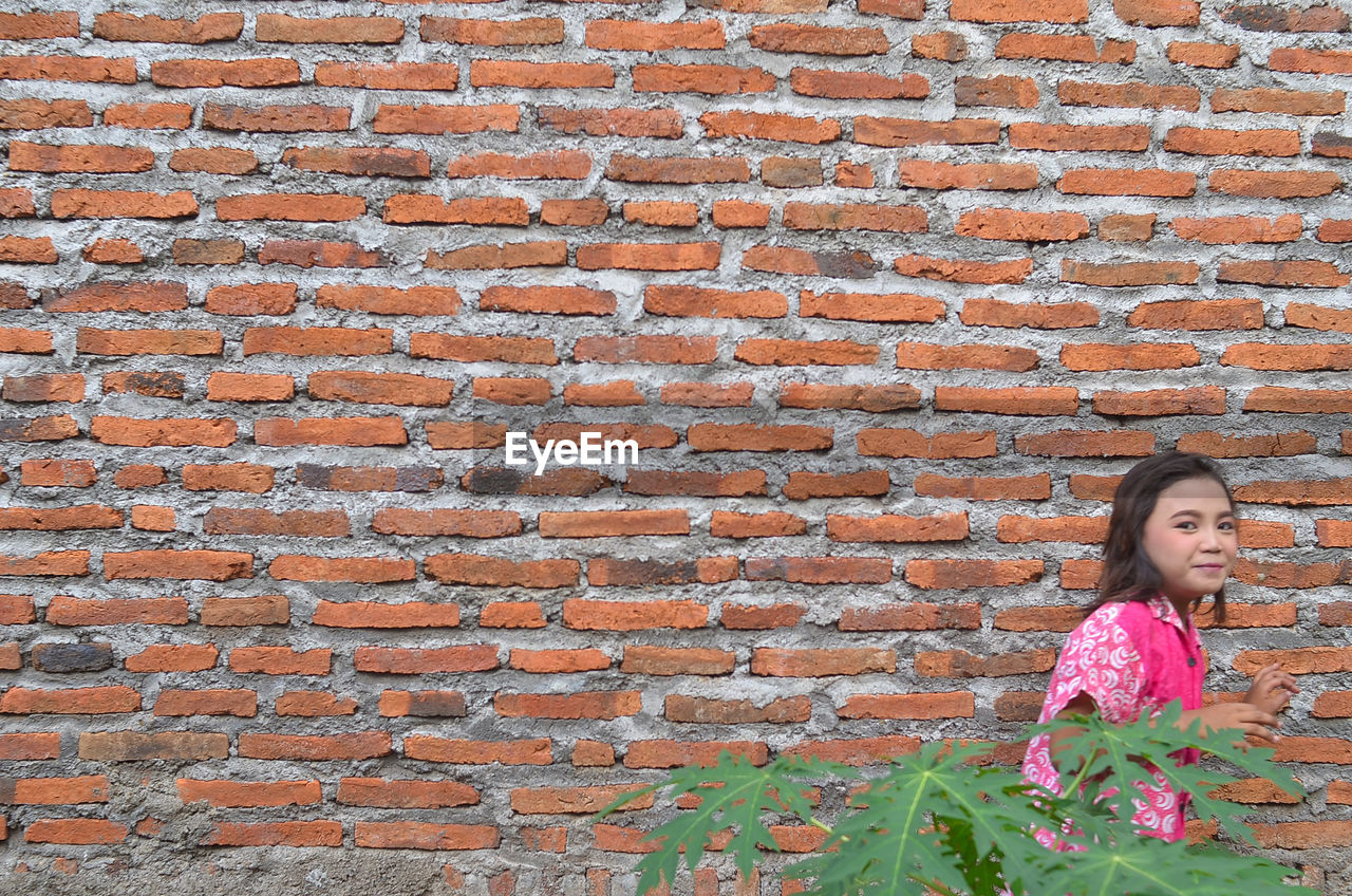 one person, brick, brick wall, wall, women, architecture, wall - building feature, child, built structure, female, standing, day, brickwork, leisure activity, childhood, casual clothing, flower, lifestyles, looking, portrait, outdoors, person, building exterior, nature, front view, pattern, clothing, human face, smiling, hairstyle, pink, emotion