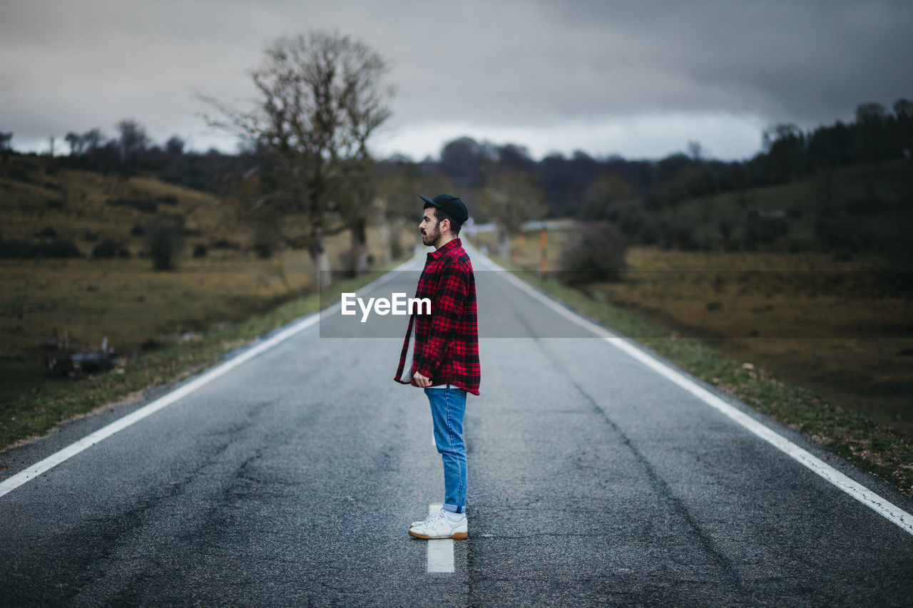 Side view of man in casual wear walking on empty asphalt road among green fields with cloudy sky on background