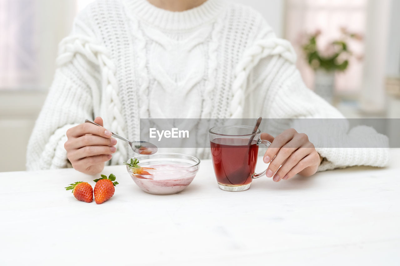 Young woman having a healthy breakfast, strawberry yogurt and tea, indoor in the kitchen. 