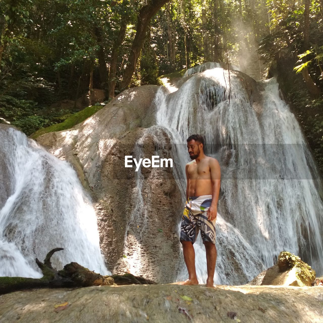 Full length of shirtless man standing against waterfall in forest